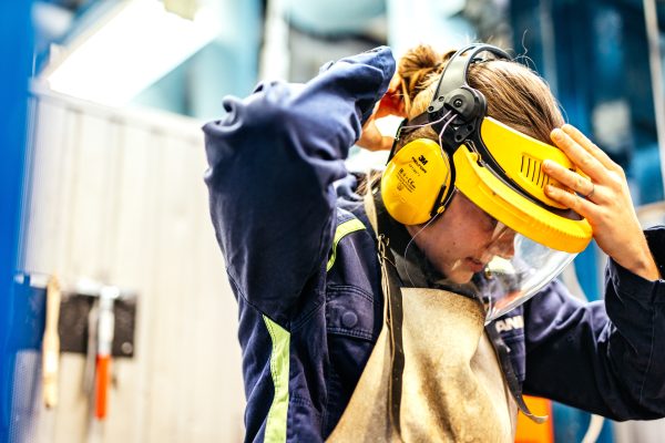 En kvinna tar på sig ett skyddsvisir med gult hörselskydd i en fabrikslokal.