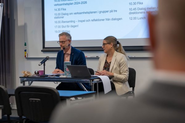 En man och en kvinna sitter vid ett bord med en stor skärm bakom sig.