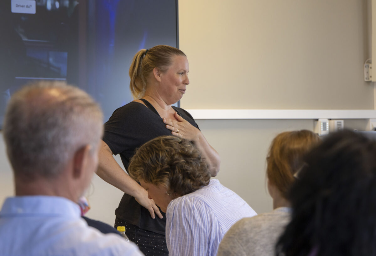 En kvinna tar sig för bröstet vid en skärm. I förgrunden flera personer som sitter och lyssnar.