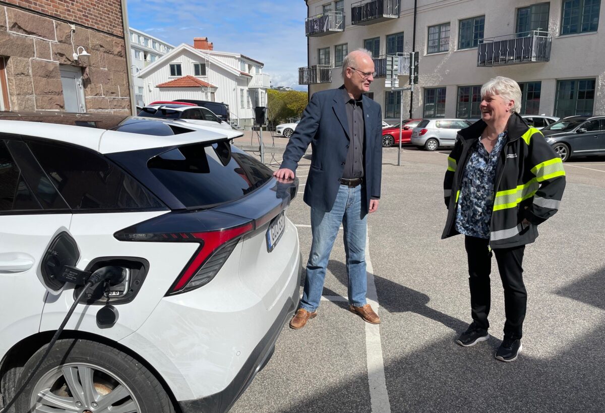 En man och en kvinna står vid en elbil som laddas. I bakgrunden hyreshus. och parkerade bilar.