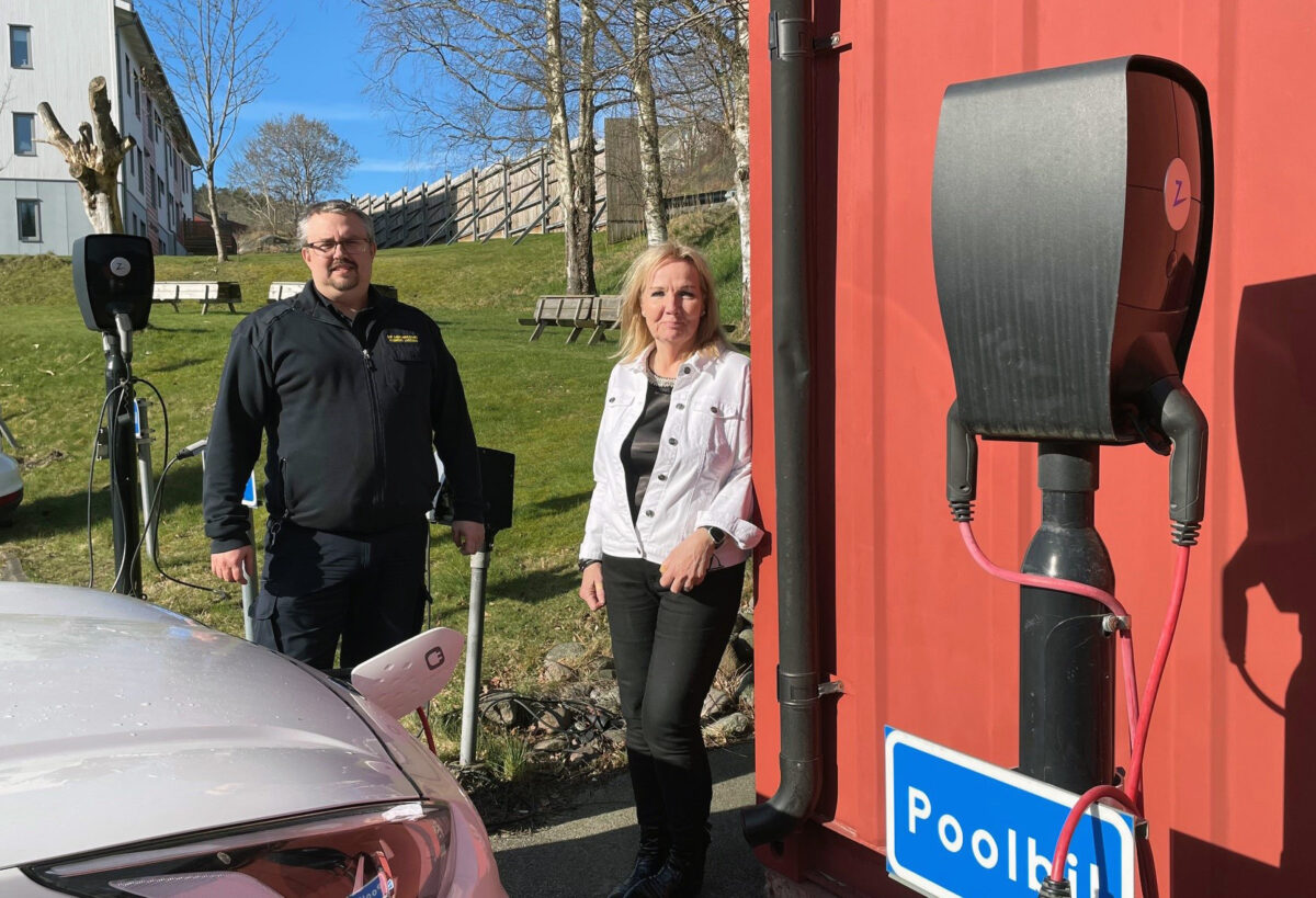 En man och en kvinna tittar in i kameran. En laddstation i förgrunden och en bil som laddas bredvid dem. Gräsmatta och träd i bakgrunden.