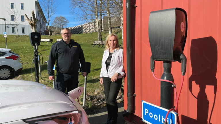 En man och en kvinna tittar in i kameran. En laddstation i förgrunden och en bil som laddas bredvid dem. Gräsmatta och träd i bakgrunden.