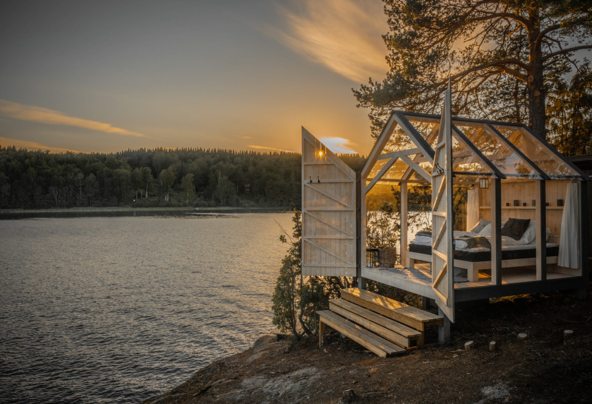 Ett växthusliknande hus med en säng och två öppna dörrar. Huset ligger vid kanten till en sjö. I bakgrunden skog och en solnedgång.