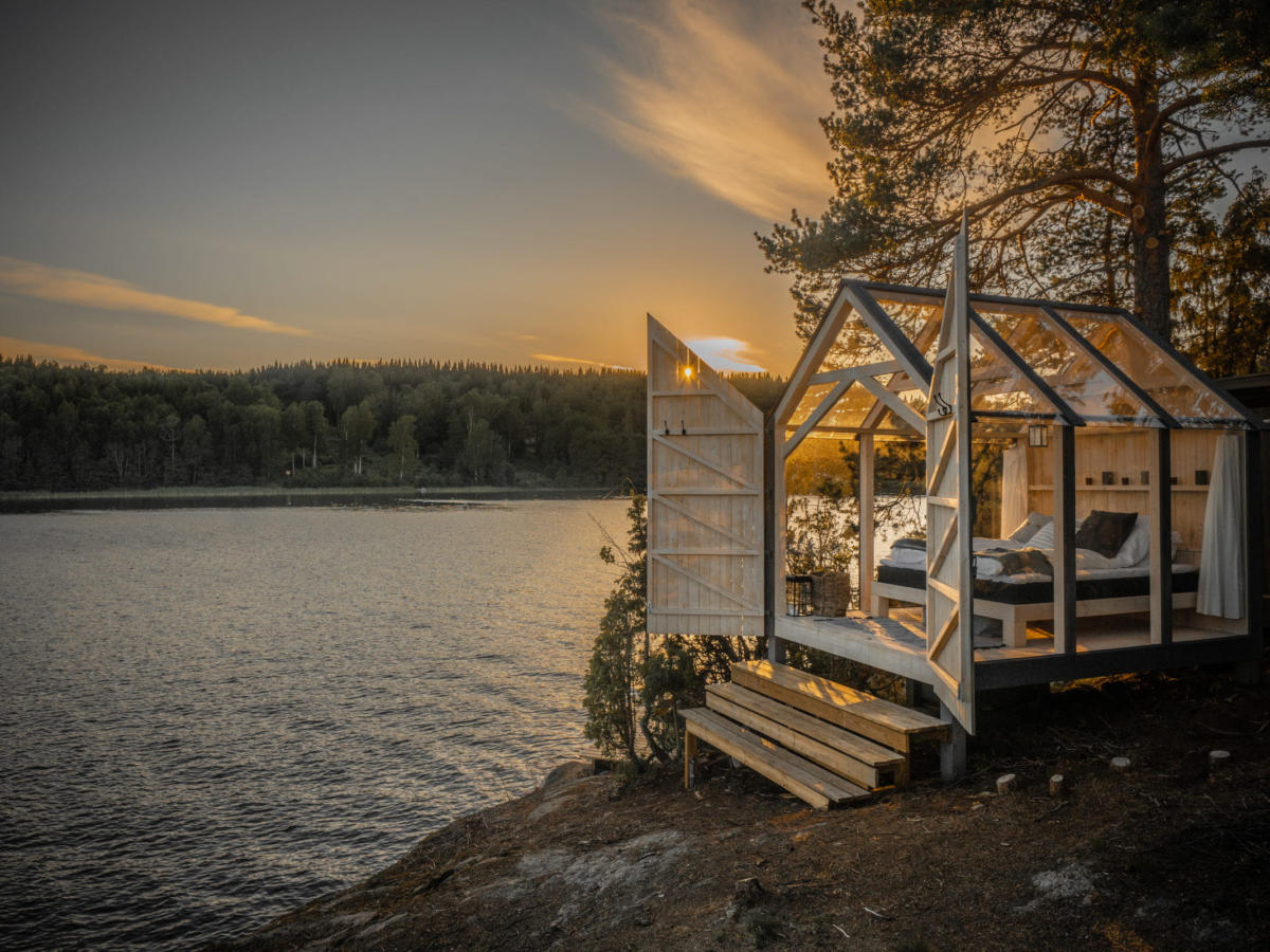 Ett växthusliknande hus med en säng och två öppna dörrar. Huset ligger vid kanten till en sjö. I bakgrunden skog och en solnedgång.