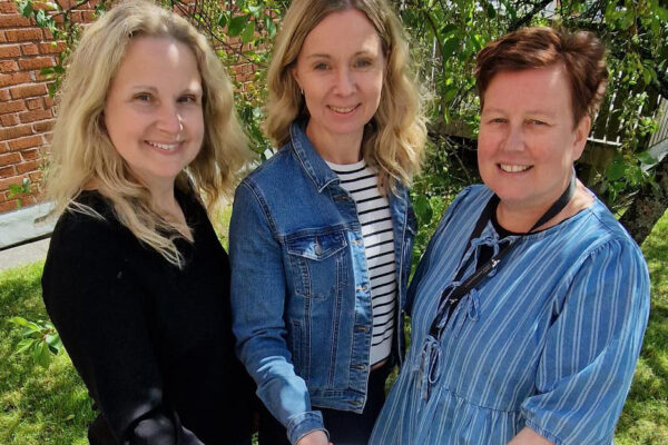 Tre kvinnor står med en framräckt dokument och ler mot kameran. Grönska i bakgrunden.