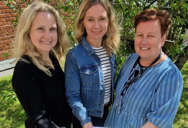 Tre kvinnor står med en framräckt dokument och ler mot kameran. Grönska i bakgrunden.