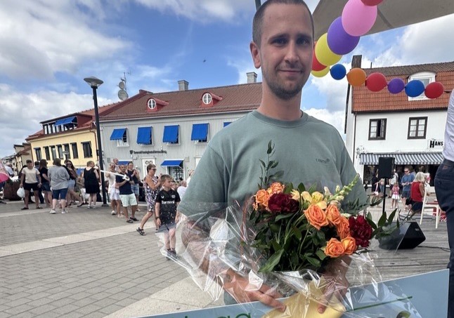 En man står med en bukett blommor. I bakgrunden ett torg med flera människor och ballonger ovanför honom.