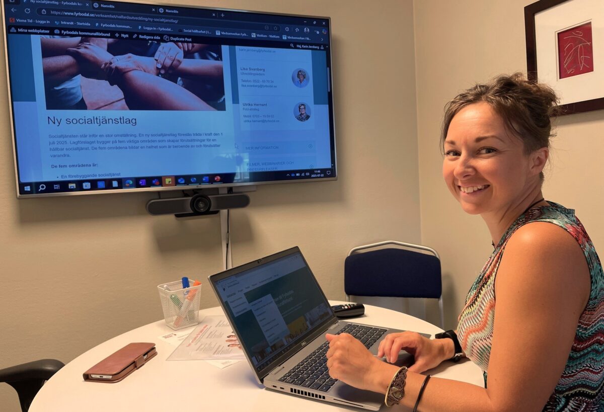 En leende kvinna sitter vid sin laptop med en stor skärm i bakgrunden. På skärmen står det "Ny socialtjänstlag".