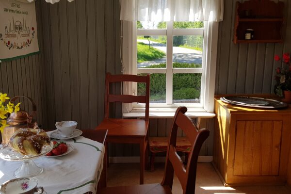 Miljöbild från en interiör av ett gammalt torp med två stolar, ett litet bord med bullar och kaffekoppar samt en bänk i ena hörnet.