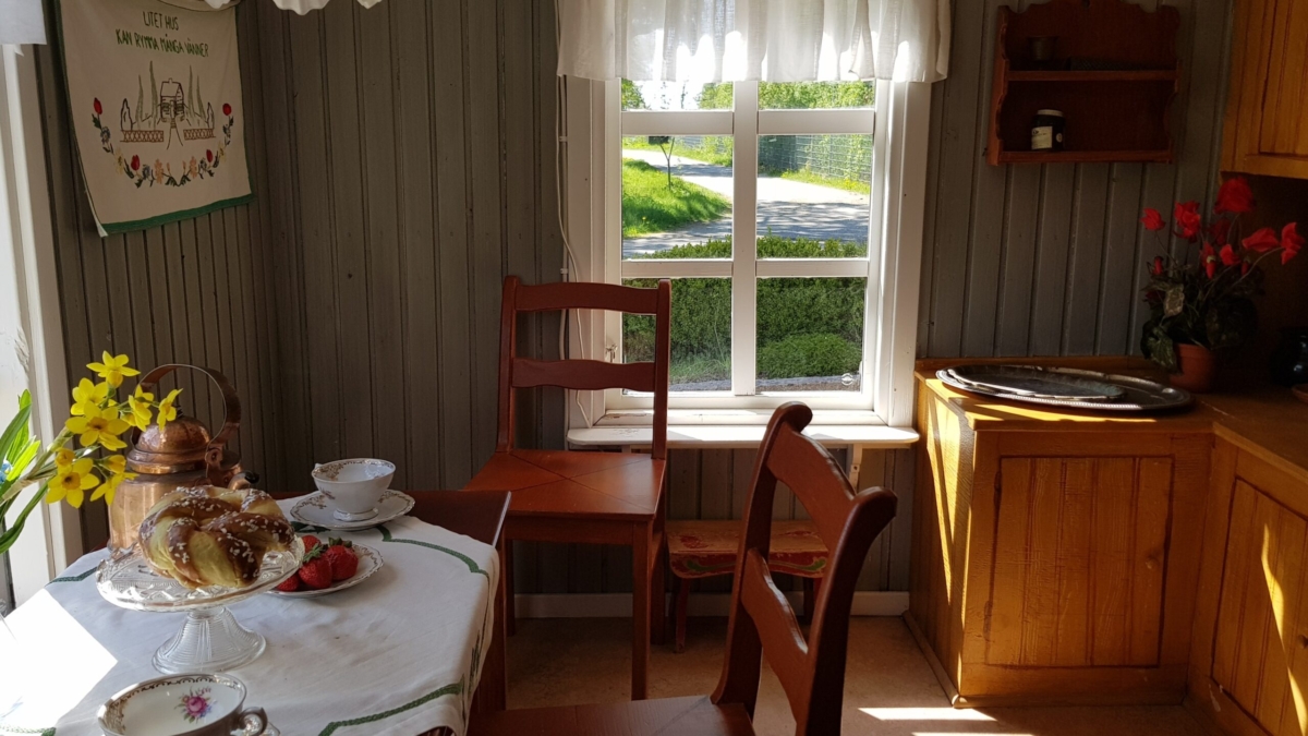 Miljöbild från en interiör av ett gammalt torp med två stolar, ett litet bord med bullar och kaffekoppar samt en bänk i ena hörnet.
