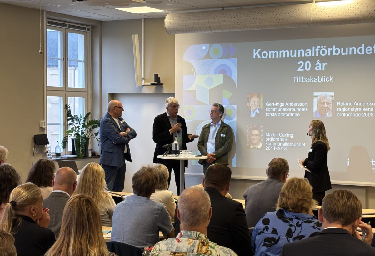 Tre personer står vid ett ståbord med en skärm i bakgrunden där det står "Kommunalförbundet 20 år". En kvinnlig moderator står vid sidan av och i förgrunden sitter flera åhörare.