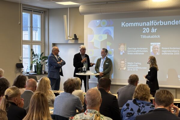 Tre personer står vid ett ståbord med en skärm i bakgrunden där det står "Kommunalförbundet 20 år". En kvinnlig moderator står vid sidan av och i förgrunden sitter flera åhörare.