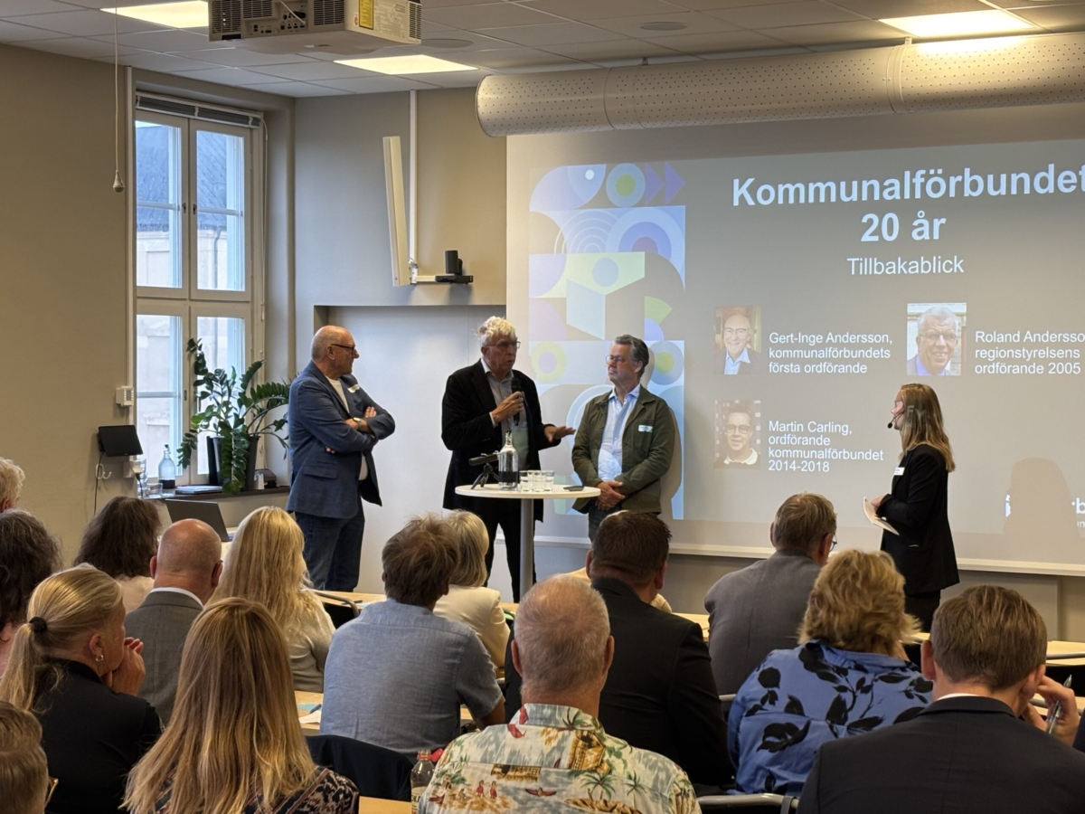 Tre personer står vid ett ståbord med en skärm i bakgrunden där det står "Kommunalförbundet 20 år". En kvinnlig moderator står vid sidan av och i förgrunden sitter flera åhörare.