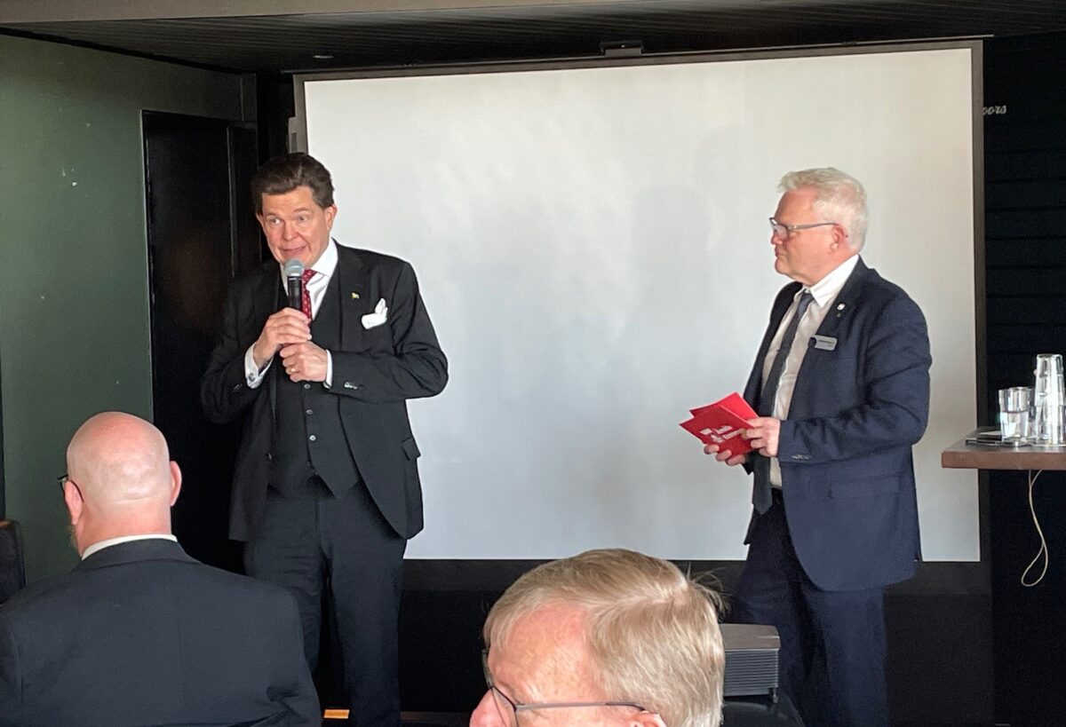 Två män står vid en vit skärm. Den ena, talman Andreas Norlén, talar i en mikrofon. Den andra står bredvid med talkort i handen.