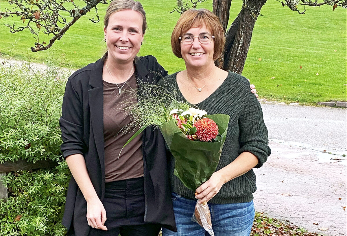 Två leende kvinnor tittar in i kameran. Kvinnan till höger håller i en blomsterbukett.I bakgrunden en gräsmatta och ett träd.