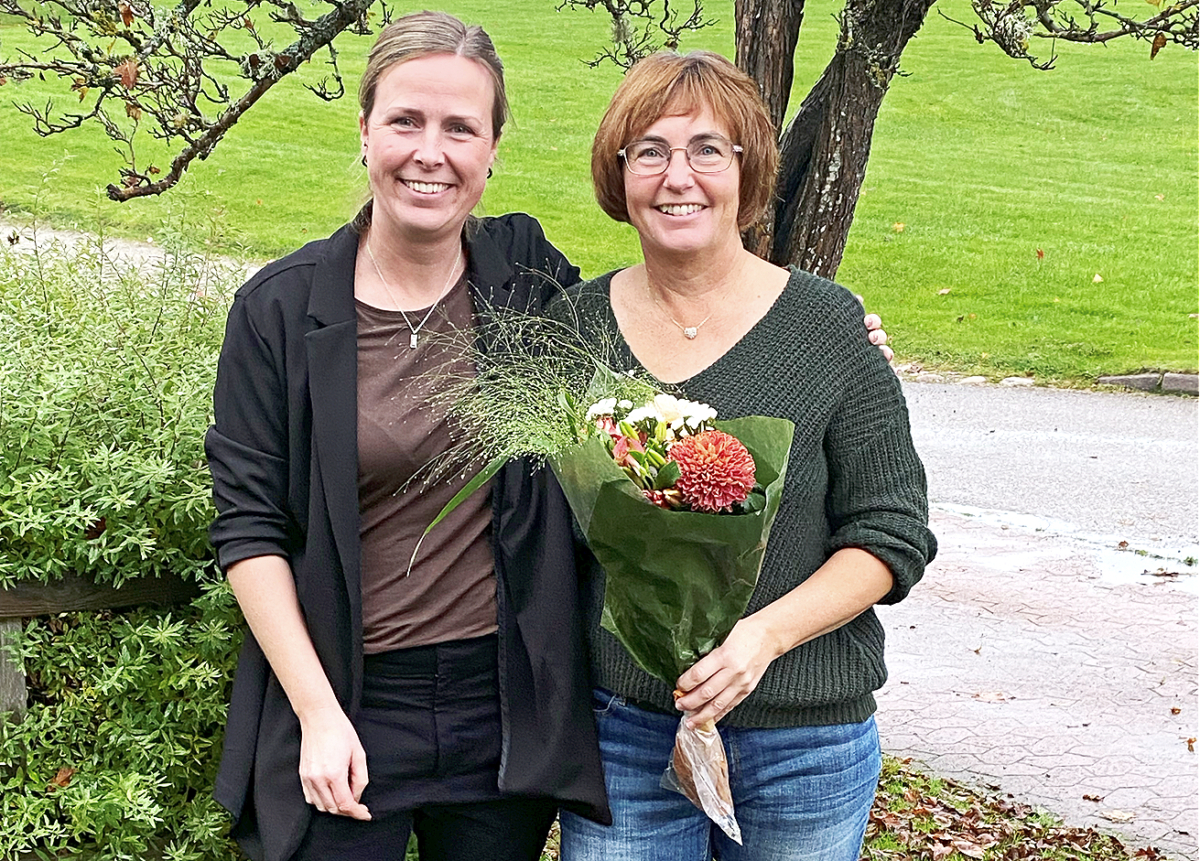Två leende kvinnor tittar in i kameran. Kvinnan till höger håller i en blomsterbukett.I bakgrunden en gräsmatta och ett träd.