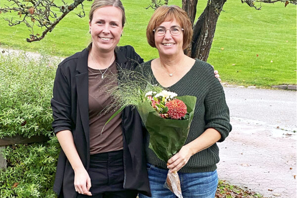 Två leende kvinnor tittar in i kameran. Kvinnan till höger håller i en blomsterbukett.I bakgrunden en gräsmatta och ett träd.