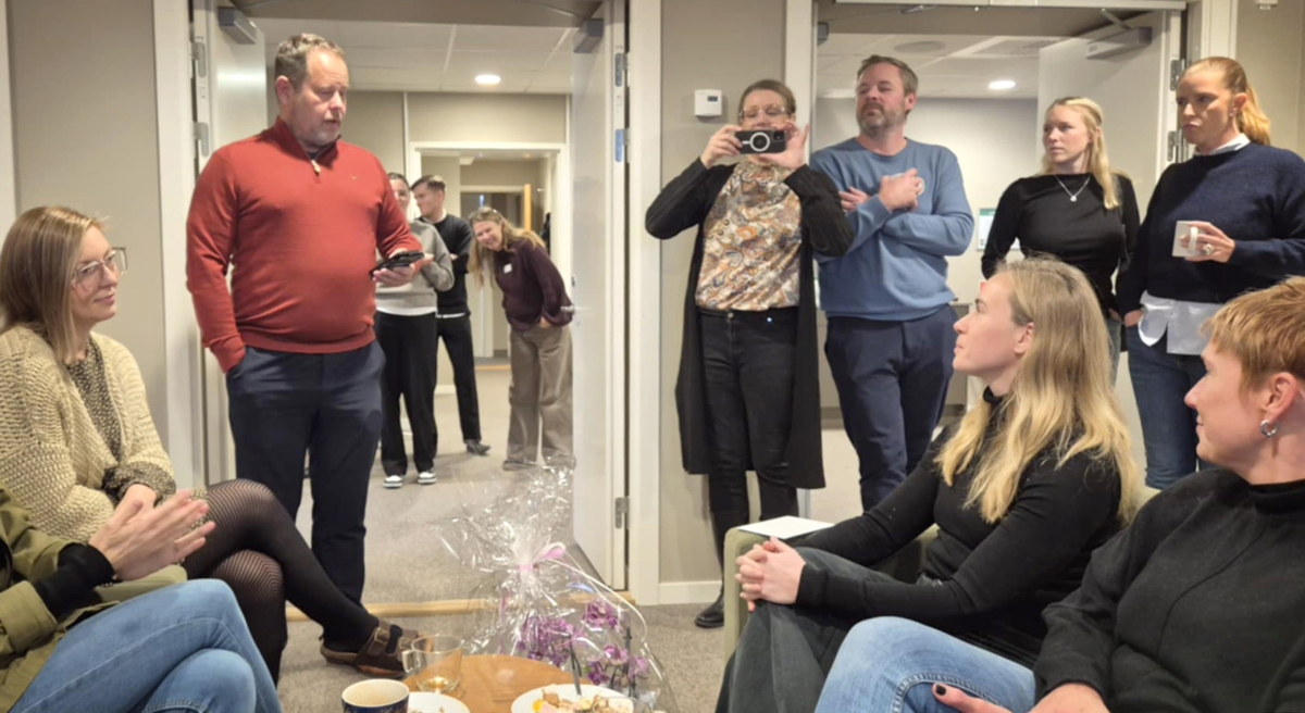 Flera personer är samlade runt en kvinna där en man håller tal samtidigt som han håller i en mobiltelefon. En kvinna i mitten tar en bild med en kamera.