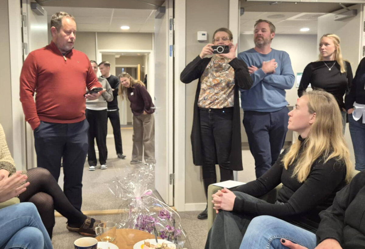 Flera personer är samlade runt en kvinna där en man håller tal samtidigt som han håller i en mobiltelefon. En kvinna i mitten tar en bild med en kamera.