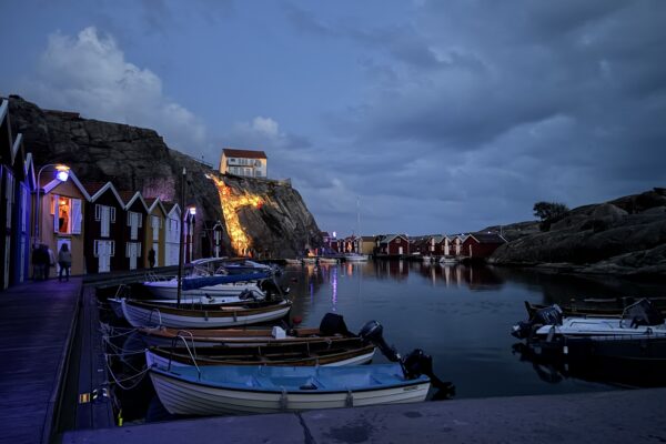 Skymningsbild vid Smögenbryggan. Flera sjöbodar i olika färger och bredvid dessa flera båtar.