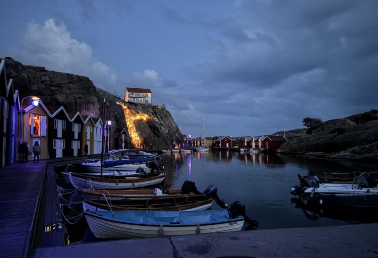 Skymningsbild vid Smögenbryggan. Flera sjöbodar i olika färger och bredvid dessa flera båtar.