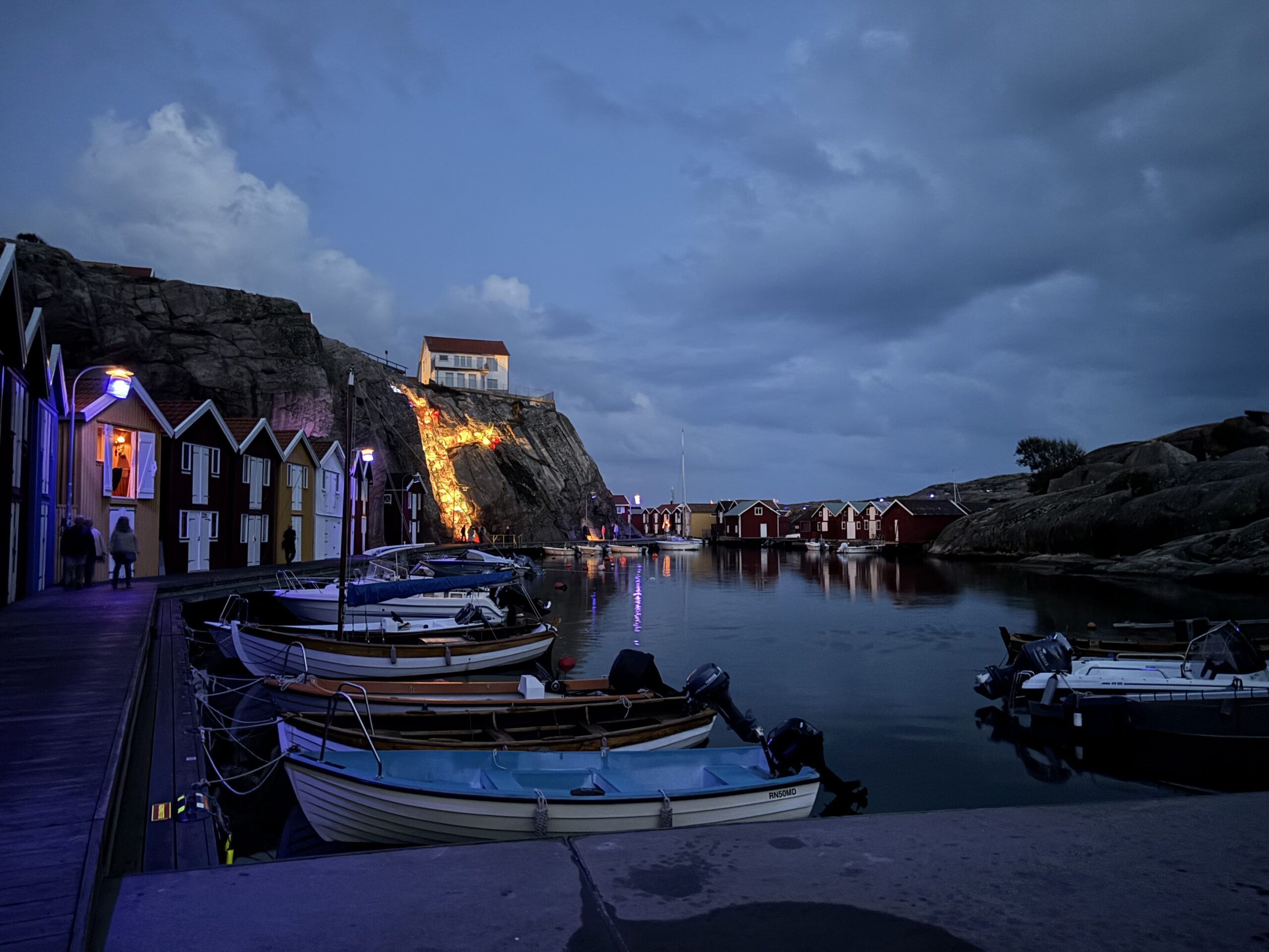 Skymningsbild vid Smögenbryggan. Flera sjöbodar i olika färger och bredvid dessa flera båtar.