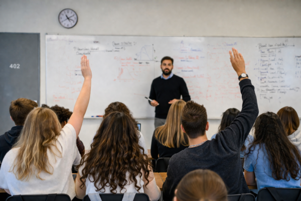 Flera ungdomar sitter i ett klassrum med ryggarna mot kameran. En lärare undervisar i förgrunden vid en whiteboard. Två av eleverna räcker upp handen.