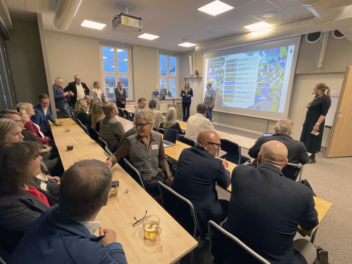 Flera rader av åhörare sitter och lyssnar på föredragshållare framför en stor skärm.
